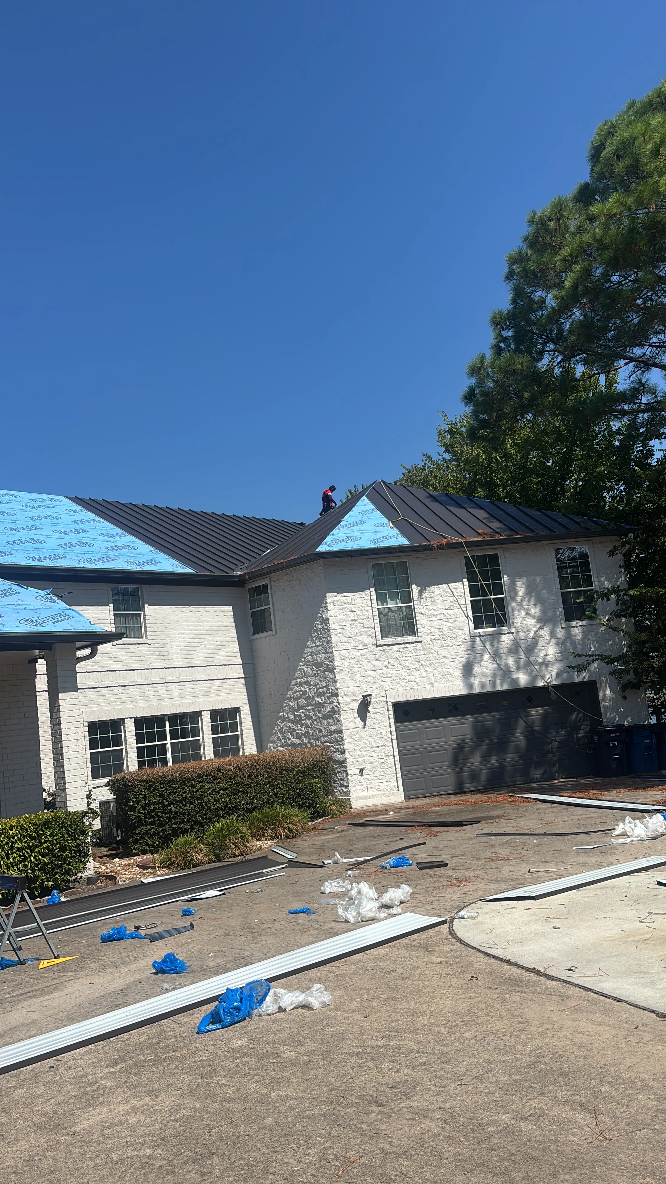 Standing seam metal roof installation on white brick two-story home with blue underlayment protection