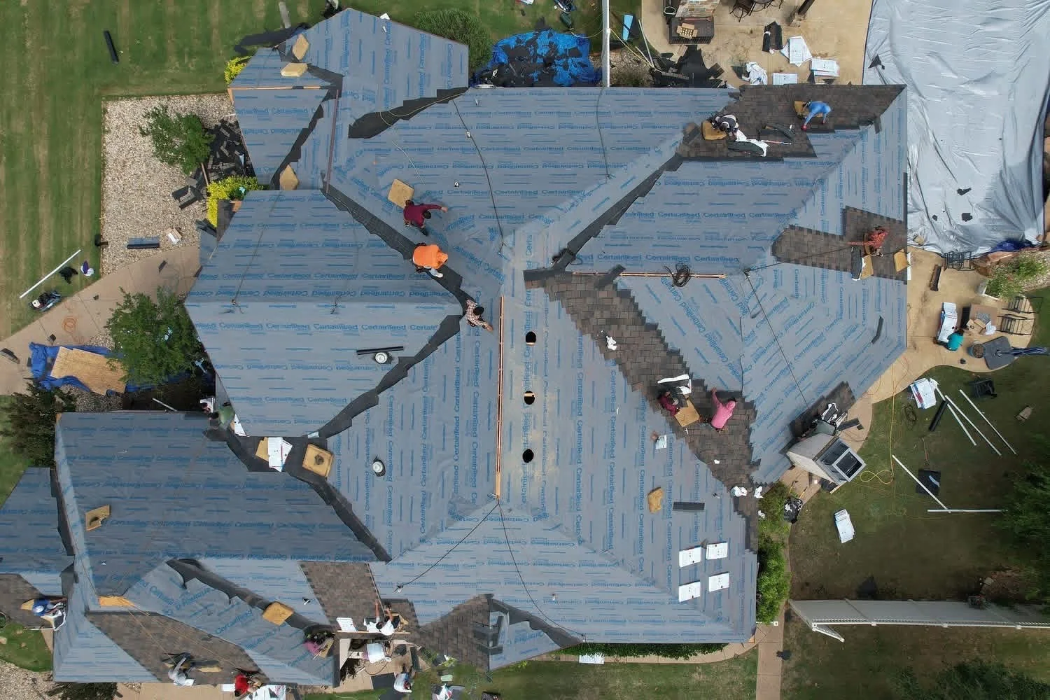 Aerial view of roof replacement with blue underlayment, multiple roof sections, and workers installing dark shingles